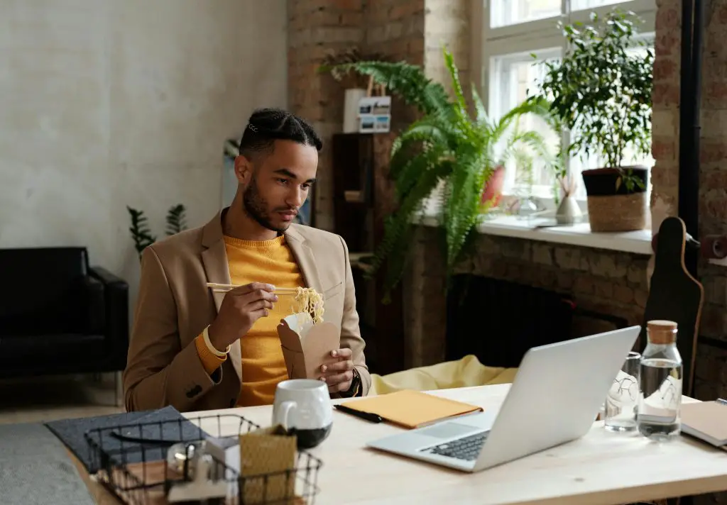 Man in brown blazer looking at a laptop while eating noodles. Photo by Tony Schnagl.